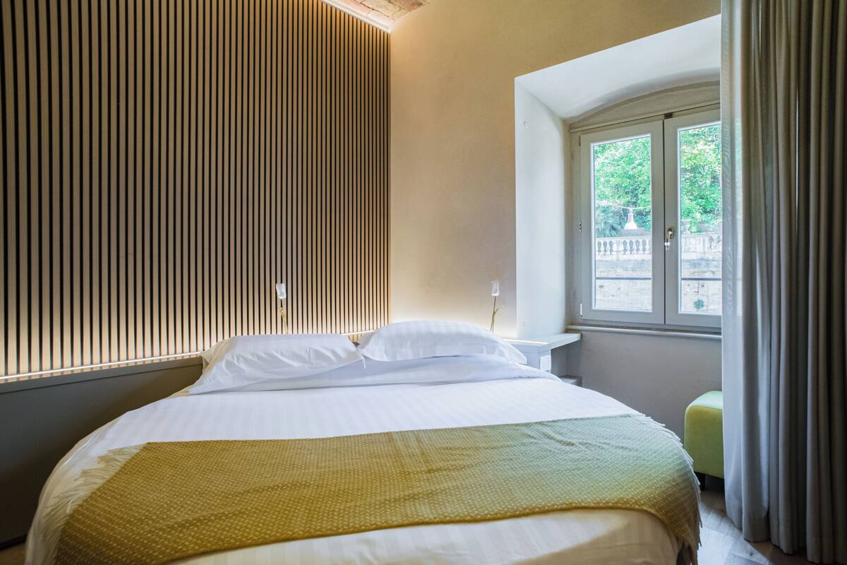 A hotel room with a bed, a bedside table, a window with curtains, and a wooden slat wall.
