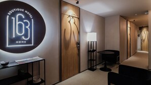Un pasillo de hotel con un letrero circular que dice 'Boutique Hotel 163 Rome', un armario de madera, una pequeña mesa con una lámpara y una silla negra.