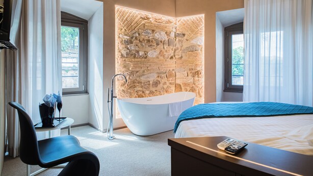 Habitación de hotel moderna con pared de detalles de piedra, una bañera, una cama con manta azul, un escritorio con control remoto y una silla.