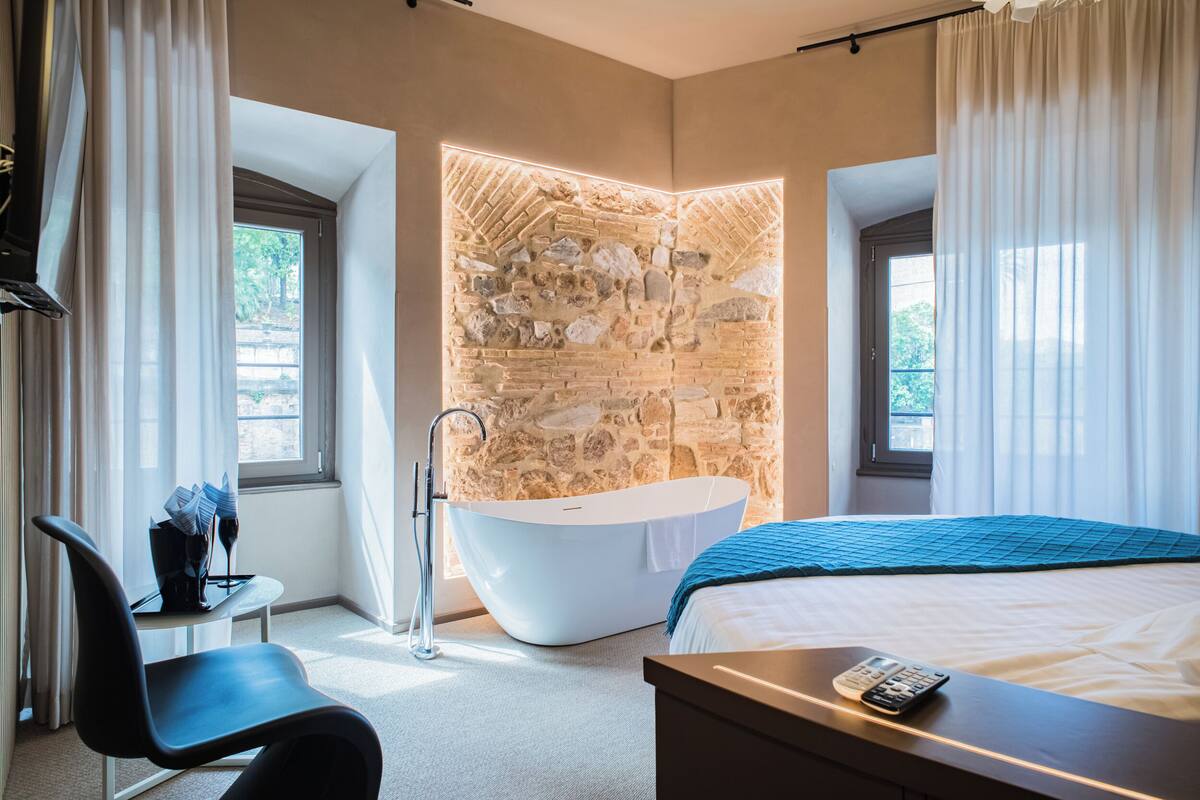 A modern hotel room with a stone accent wall, a bathtub, a bed with a blue blanket, a desk with a remote control, and a chair.