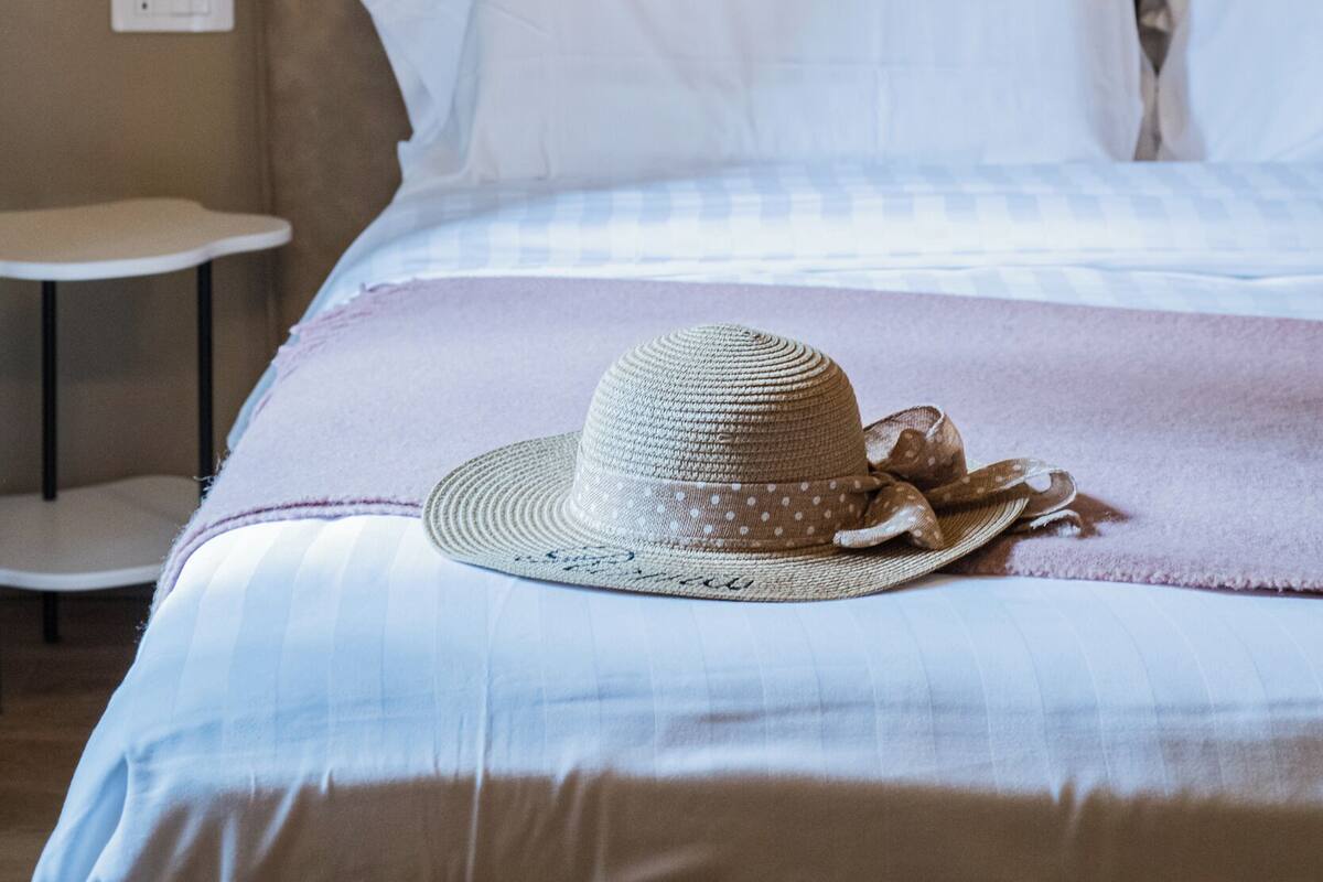 A neatly made bed with white linens, a round straw hat with a ribbon, and a small round table beside it.