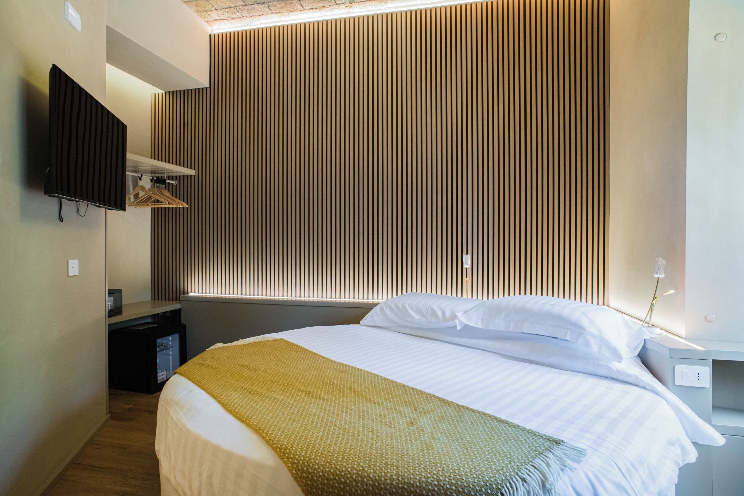 A modern hotel room with a bed, a TV mounted on the wall, and a wooden slat headboard.