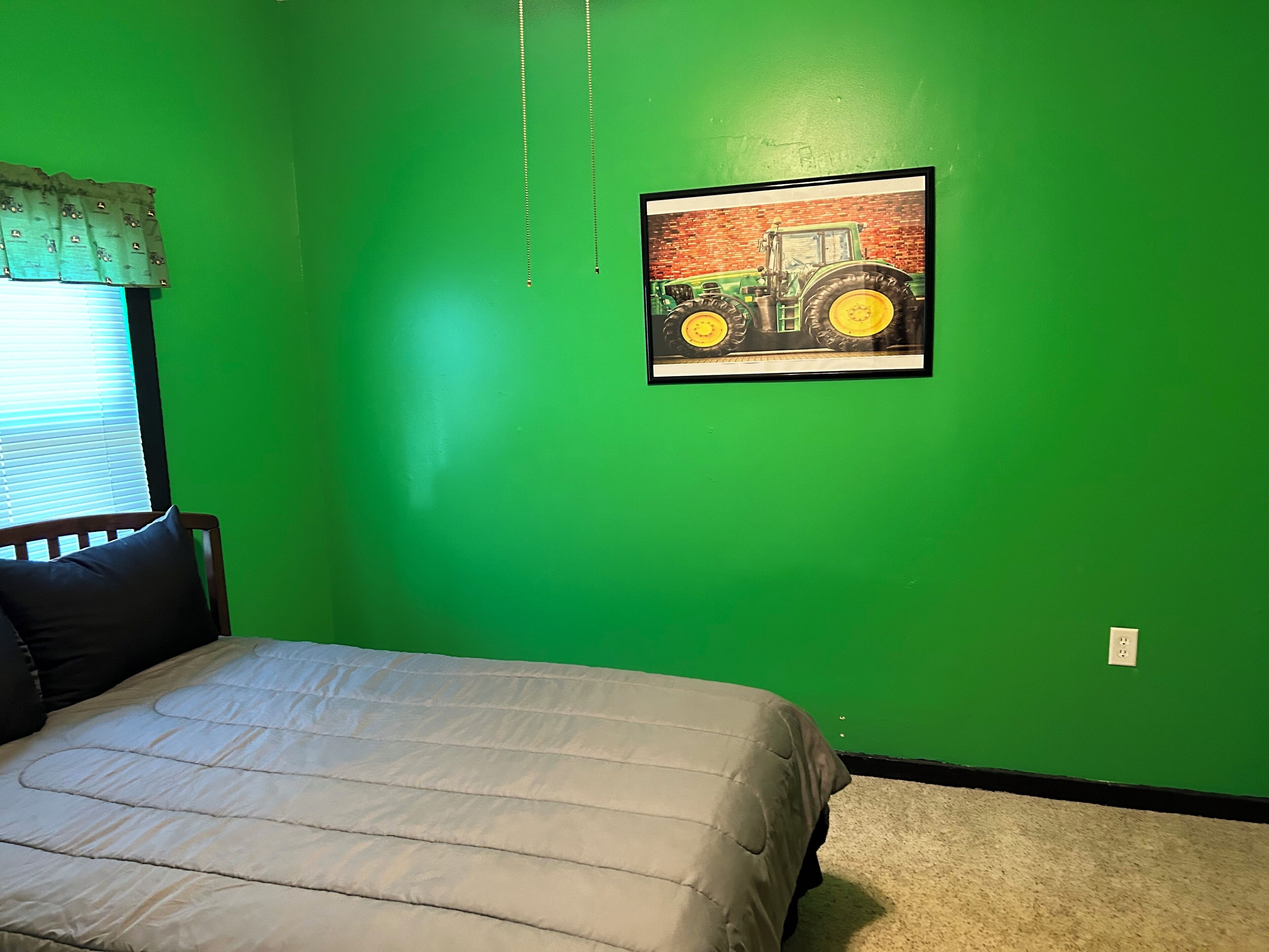 A bedroom with a green wall, a framed picture of a tractor, a bed with a gray blanket, and a window with blinds.