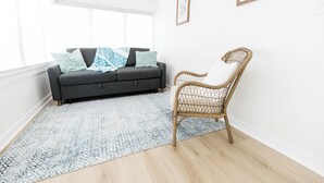 A room with a grey sofa, a wicker chair, and a patterned rug. - Best Value 2BR Midtown Stay - Clean + Central (Omaha)