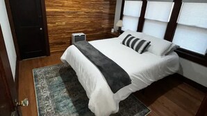 A bedroom with a wooden accent wall, a bed with white bedding, a patterned rug, and a window with blinds. - Best Value 2BR Midtown Stay - Clean + Central (Omaha)