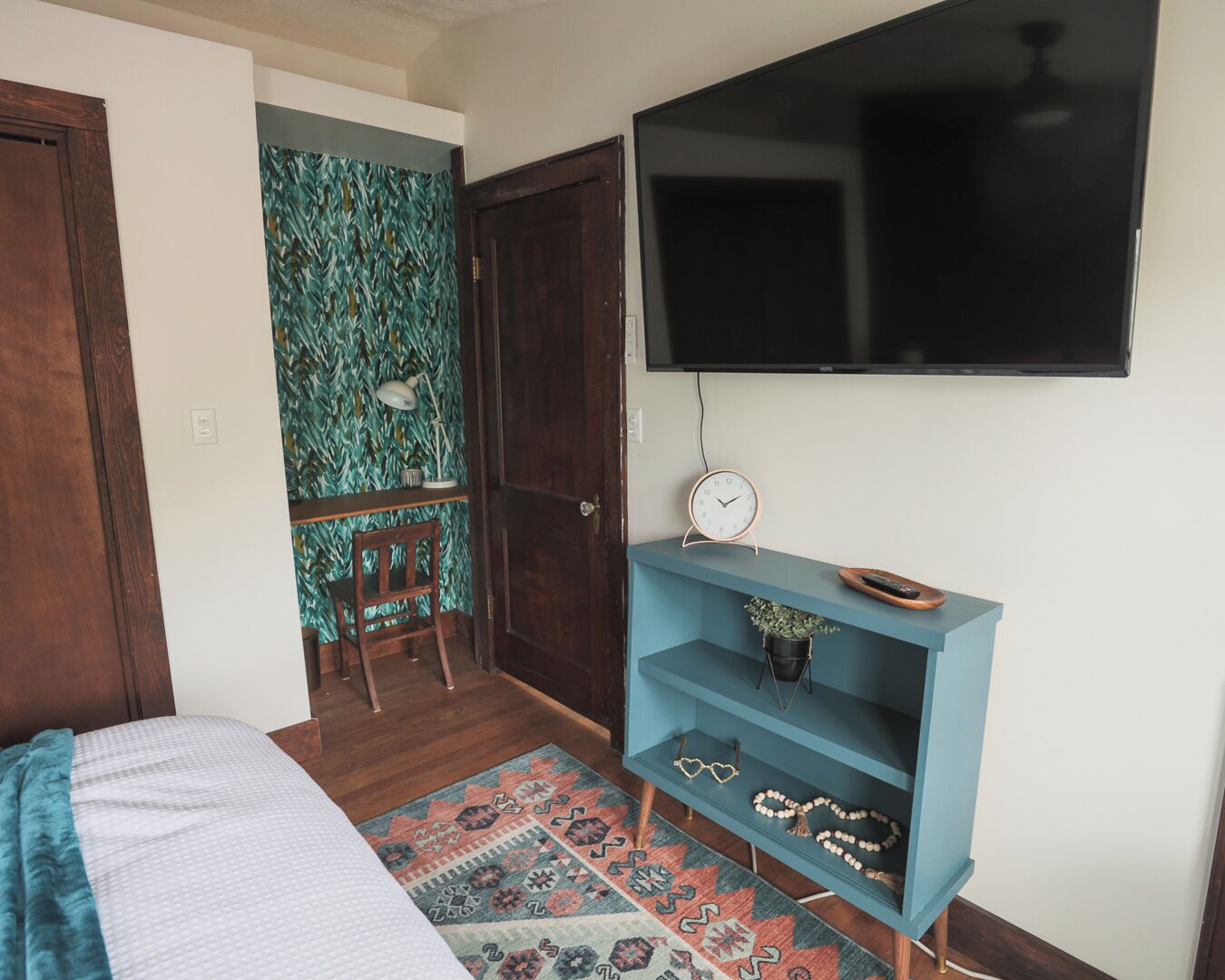 Ein Schlafzimmer mit einem Bett, einem an der Wand montierten Fernseher, einem blauen Regal mit Dekoration und einem gemusterten Teppich.