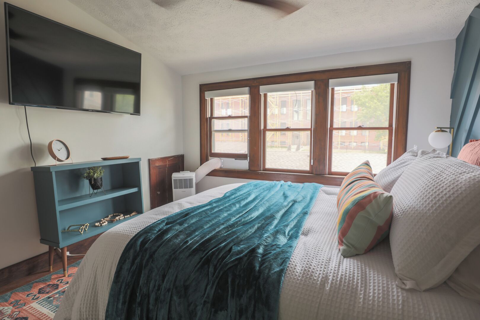 A bedroom with a bed, a TV mounted on the wall, a window, and a desk with a chair.