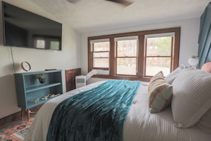 A bedroom with a bed, a TV mounted on the wall, a window, and a desk with a chair.