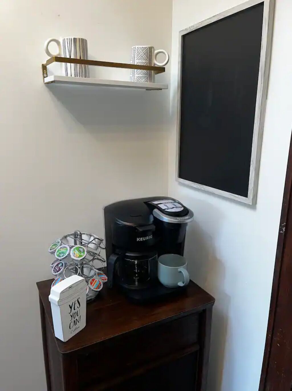 Een koffiezetapparaat op een houten tafel met een mok, een houder voor koffiekapsules, en een bordje met de tekst 'YES YOU CAN! Coffee'.