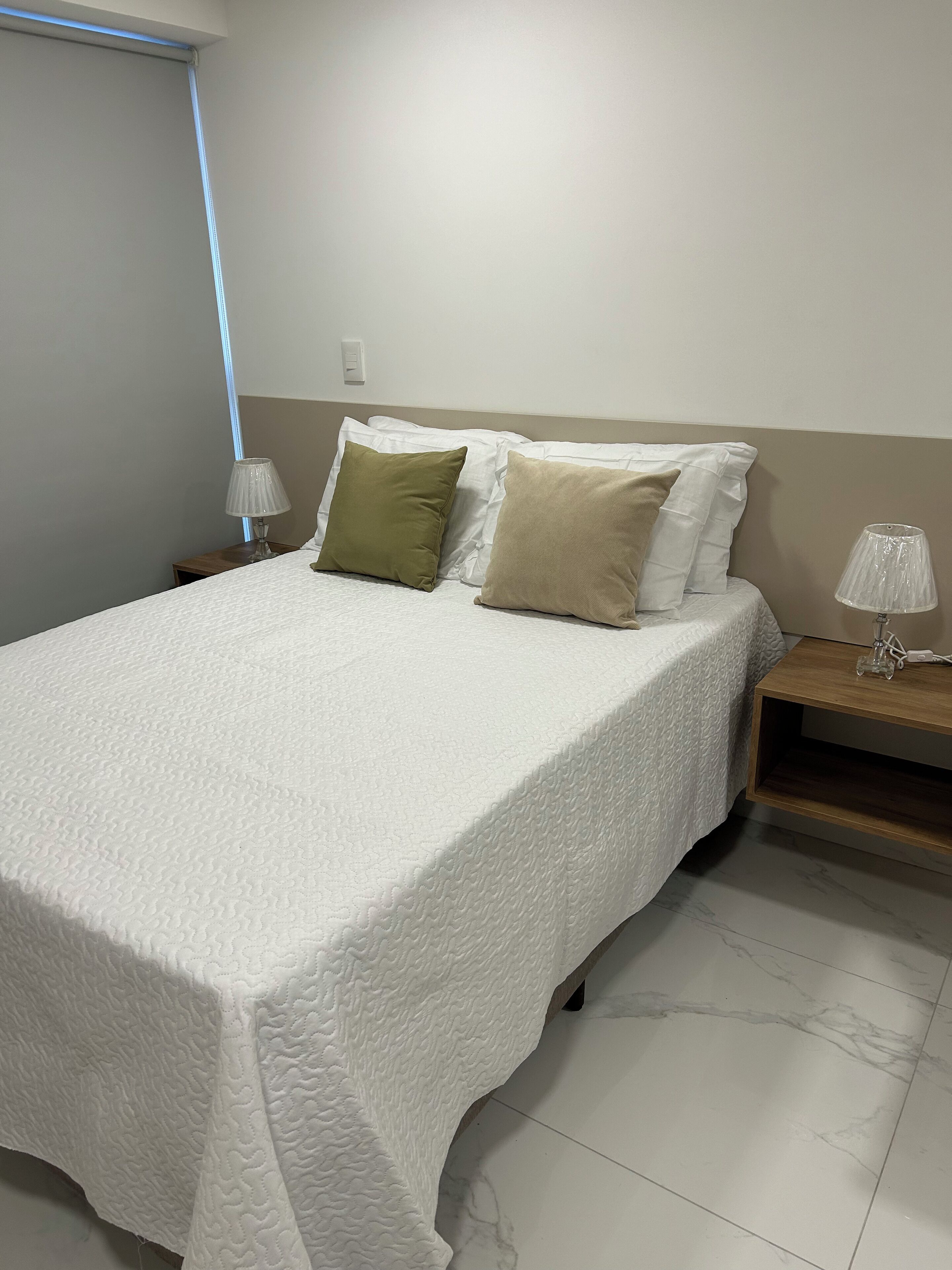 Una cama bien tendida con un edredón blanco, dos almohadas de colores diferentes y dos lámparas de noche sobre mesitas de madera.