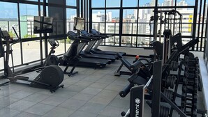 A gym with various exercise equipment, large windows, and a city view. - Melhores Flats - Perfeição perto do mar (João Pessoa)