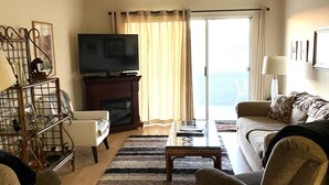 A living room with a TV, two armchairs, a sofa, a coffee table, and a view through a glass door.