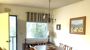 A dining area with a round wooden table, six chairs, a chandelier, a window with curtains, a bookshelf, and a framed painting on the wall.