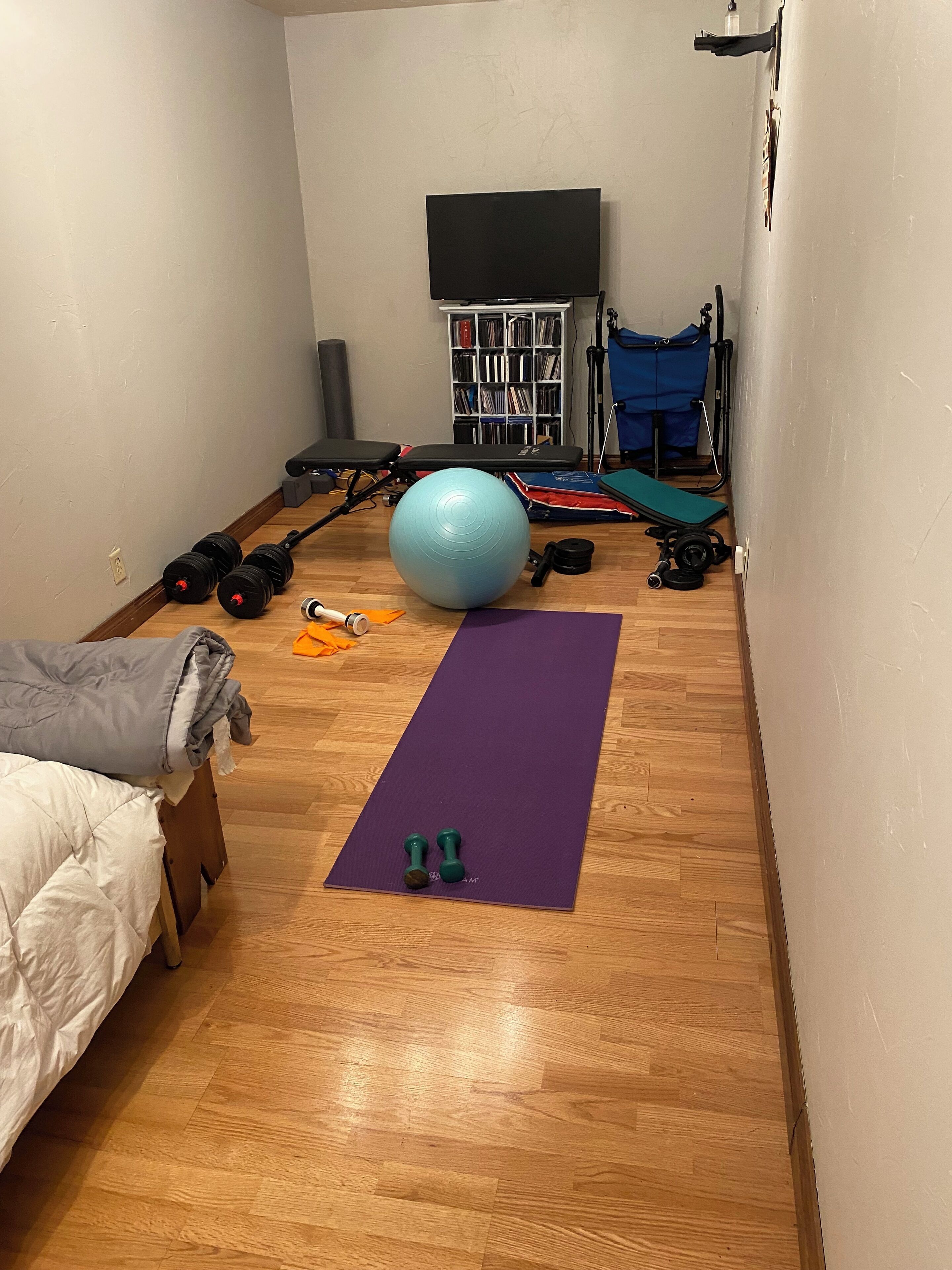 Ein Zimmer mit Holzboden, einer violetten Trainingsmatte, zwei Kurzhanteln, einem blauen Trainingsball, einer Bank und einem Bücherregal mit DVDs.