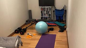 Ein Zimmer mit Holzboden, einer violetten Trainingsmatte, zwei Kurzhanteln, einem blauen Trainingsball, einer Bank und einem Bücherregal mit DVDs.