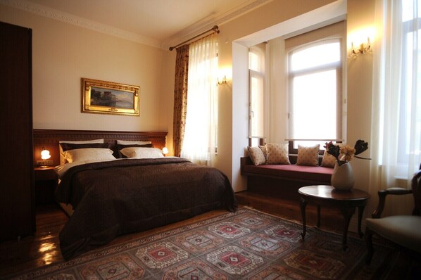 Une chambre d’hôtel avec un grand lit, un coin salon avec un banc et des coussins, une petite table avec un vase et un tableau au mur.
