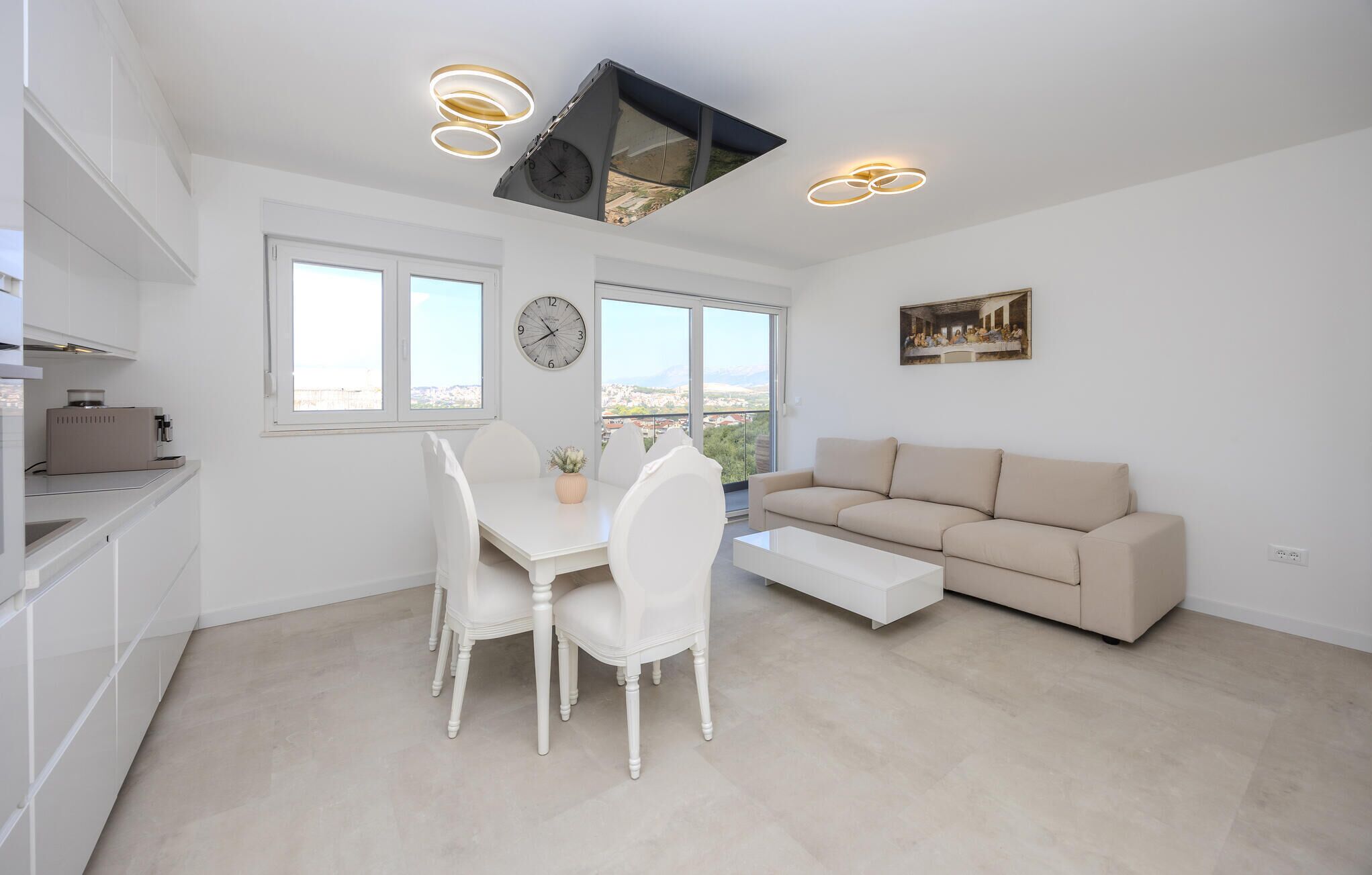 A modern kitchen with white cabinetry, a dining area with a white table and chairs, a beige sofa, and a large window offering a city view.