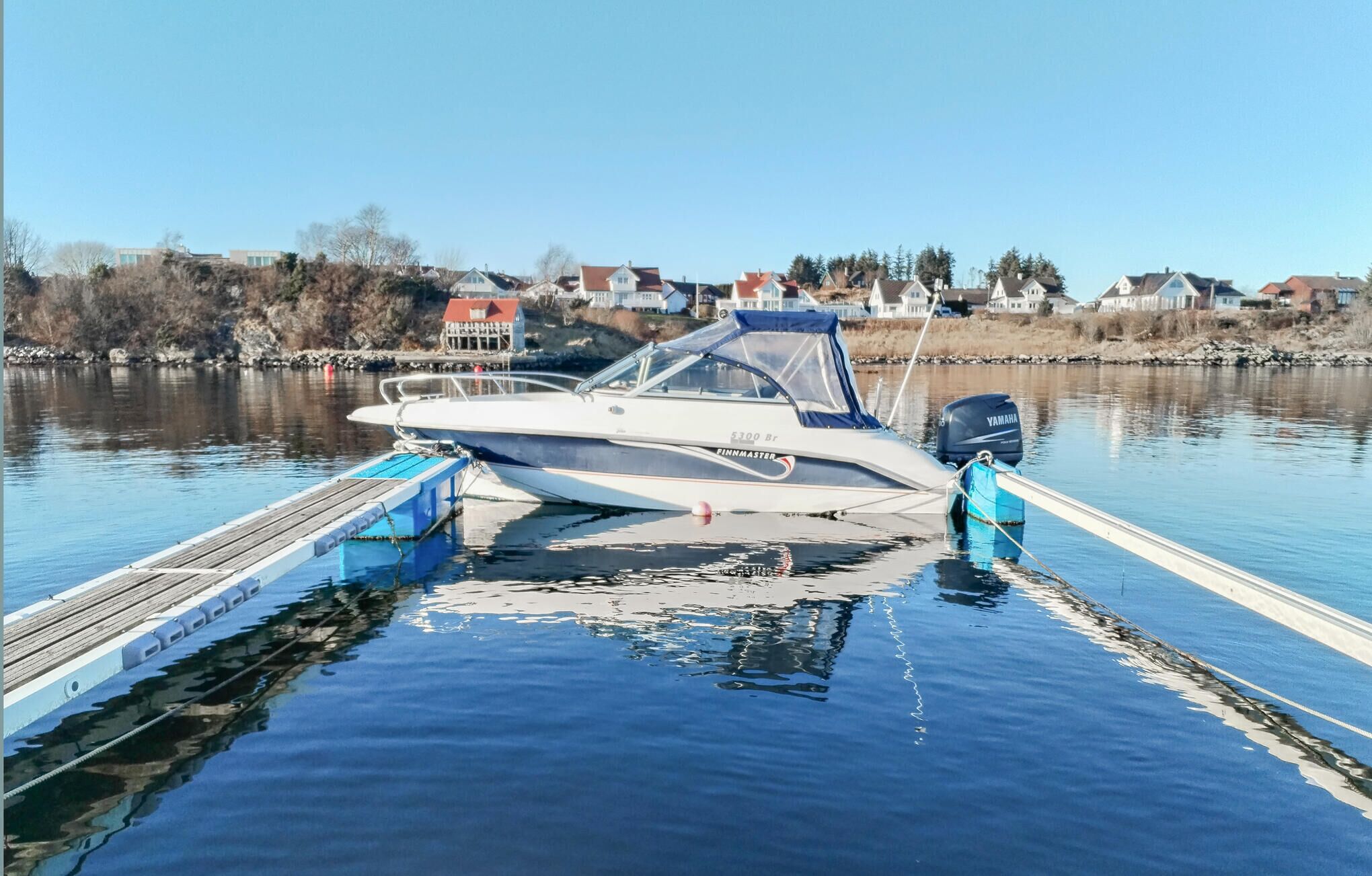 A boat docked at a pier with a calm lake and houses in the background.