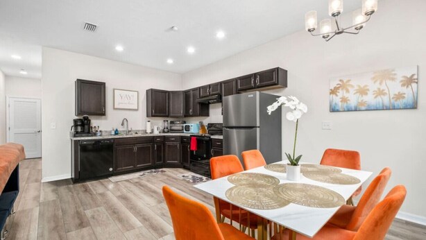 A modern kitchen with dark cabinets, a dining area with orange chairs, and a white table. - Beautiful Home Walk to UF  Newly built (Gainesville)