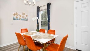 A dining area with a white table, orange chairs, a chandelier, and a window with curtains. - Beautiful Home Walk to UF  Newly built (Gainesville)