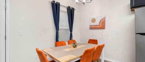 Un comedor con mesa de madera y sillas naranjas, una araña y una ventana con cortinas.