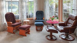 A sunlit room with a large window, a blue armchair, a wooden rocking chair, a small round table with a red tablecloth, and a brown leather chair.