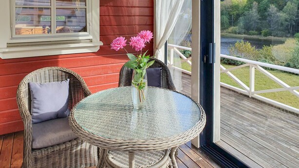 A wicker table with a glass top, two wicker chairs with cushions, and a vase with flowers on a balcony with a view of a lake and trees.