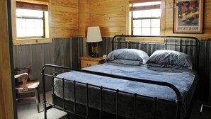 Una camera in una baita di legno con un letto in metallo, una sedia, un comodino con una lampada e un poster appeso al muro.