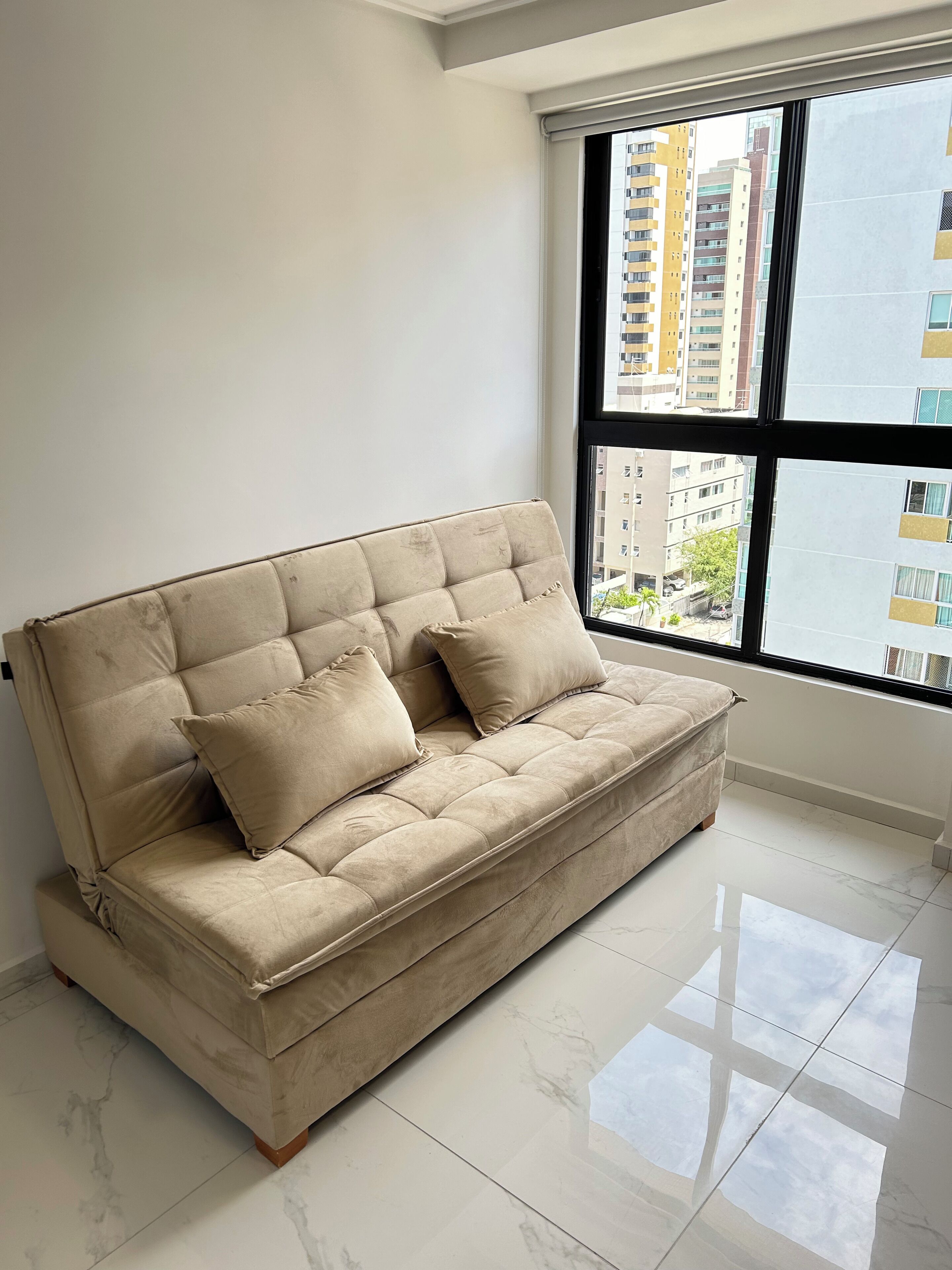A beige sofa with two cushions in a room with a large window, white walls, and a tiled floor.