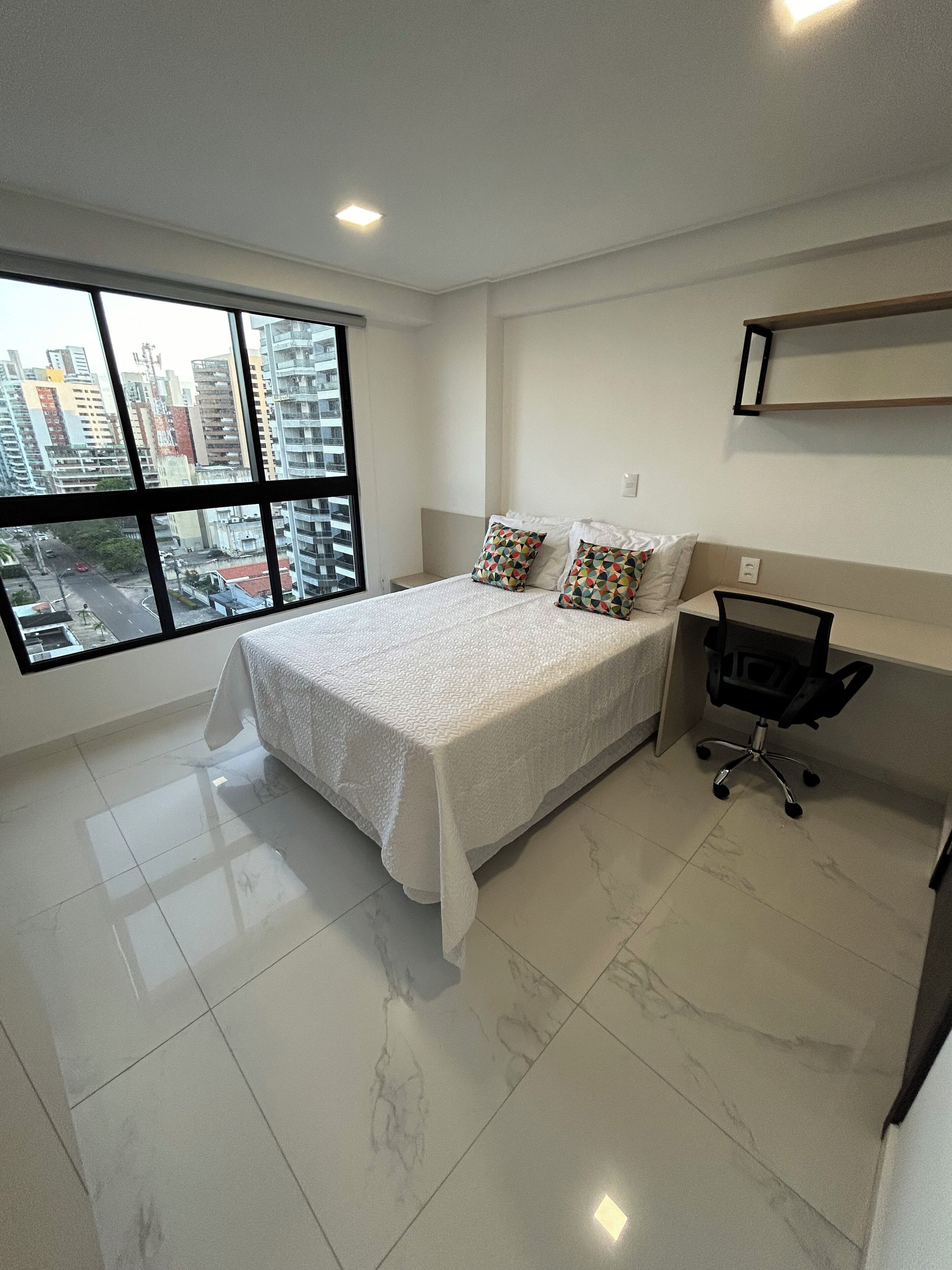 A hotel room with a large bed, a desk, and a chair. There is a window with a city view.