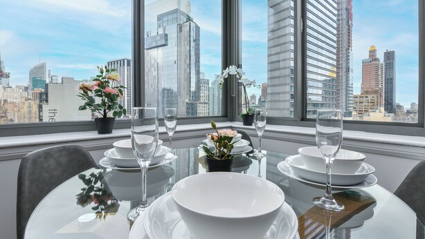Una mesa de comedor dispuesta con platos, cuencos y copas de vino, con vista a un paisaje urbano a través de grandes ventanales.