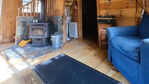 Interior de una cabaña de madera con estufa, un sofá azul, una escalera y un ventilador.