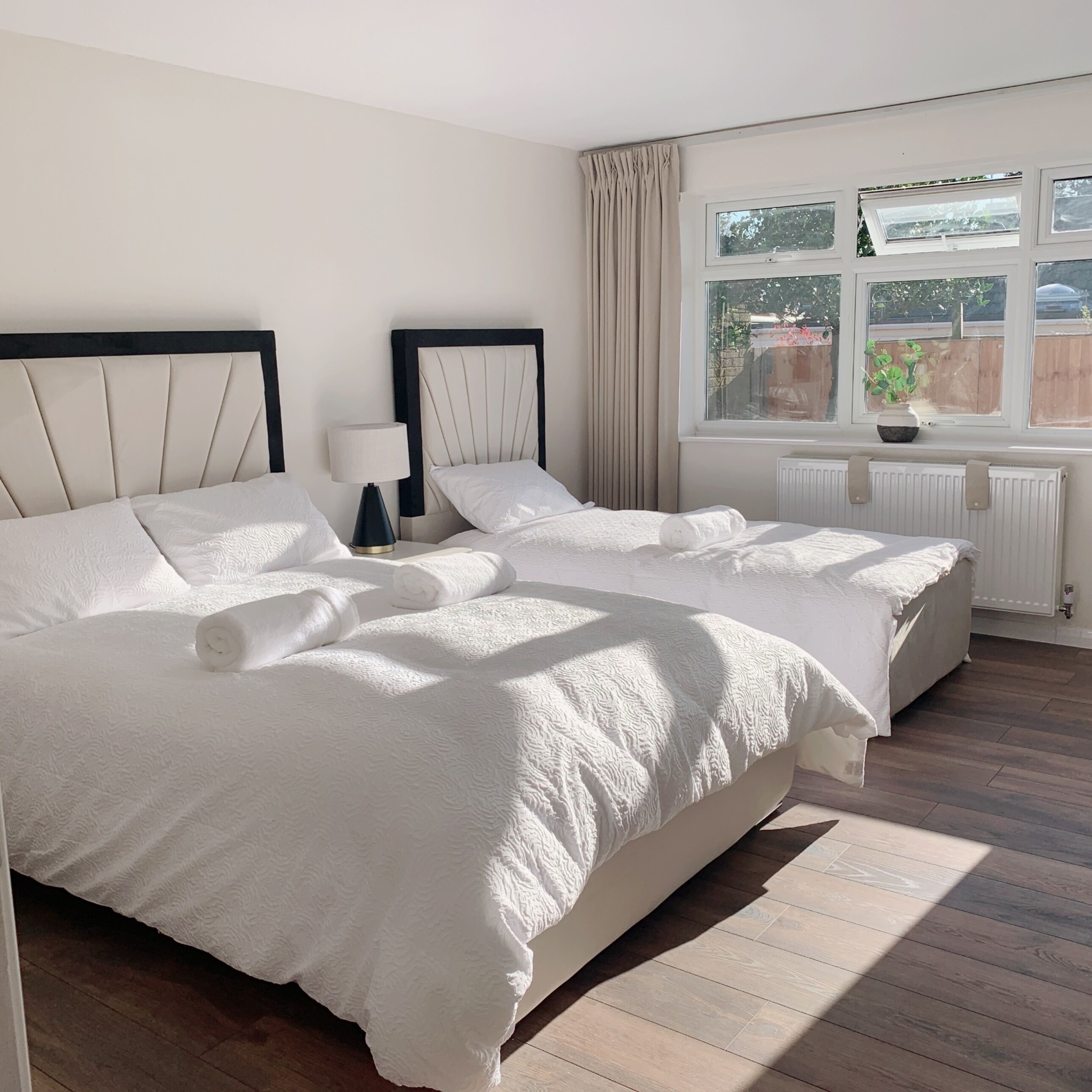 A hotel room with two beds, a window with curtains, a radiator, and a potted plant.