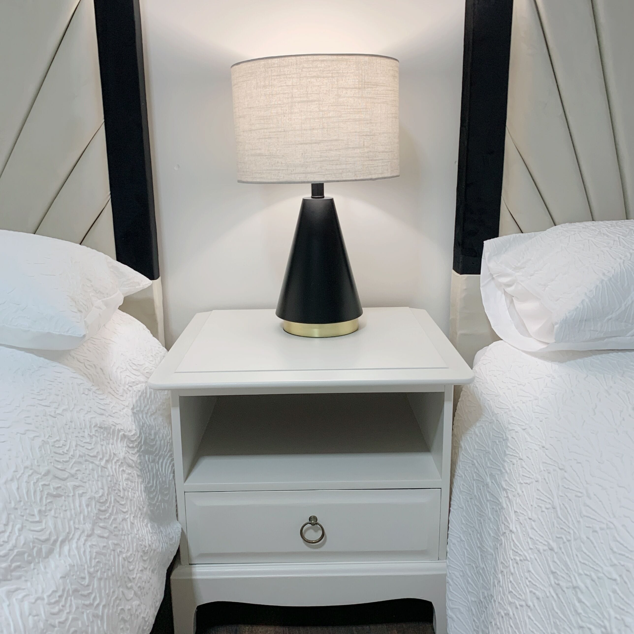 A white bedside table with a black lamp, a drawer, and two white pillows.