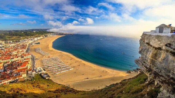 Un pueblo costero con una playa, un acantilado con un edificio en la cima y vistas al mar.
