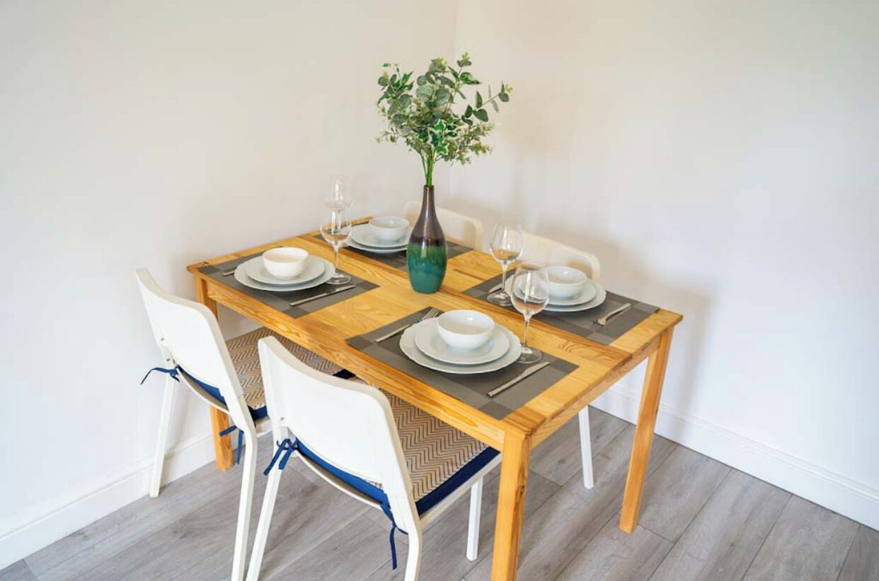 A dining table set with white plates, silverware, and wine glasses, accompanied by a vase with green foliage.