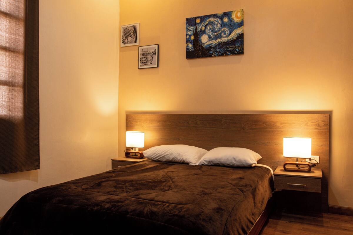 A neatly made bed with a wooden headboard, two bedside lamps, and a painting on the wall.