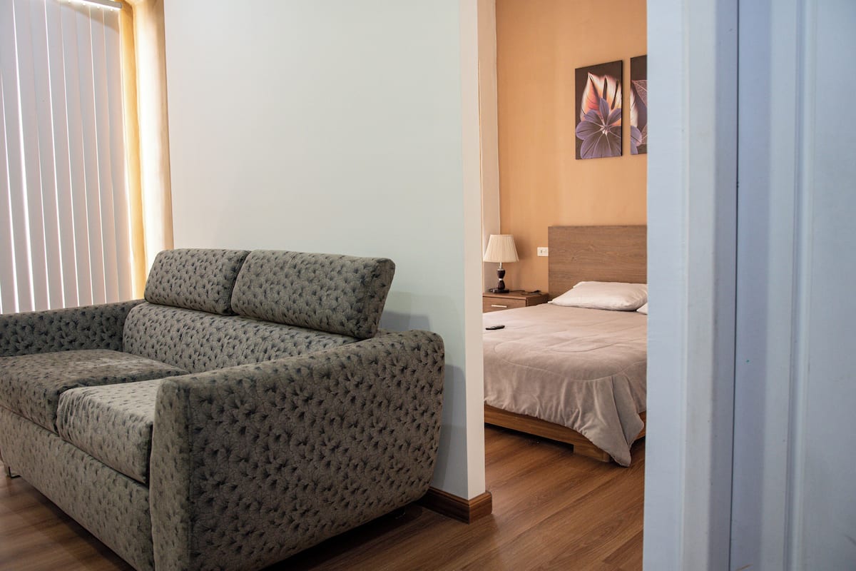 A room with a grey upholstered sofa, a wooden floor, a bed with a beige bedspread, a nightstand with a lamp, and two wall-mounted paintings.