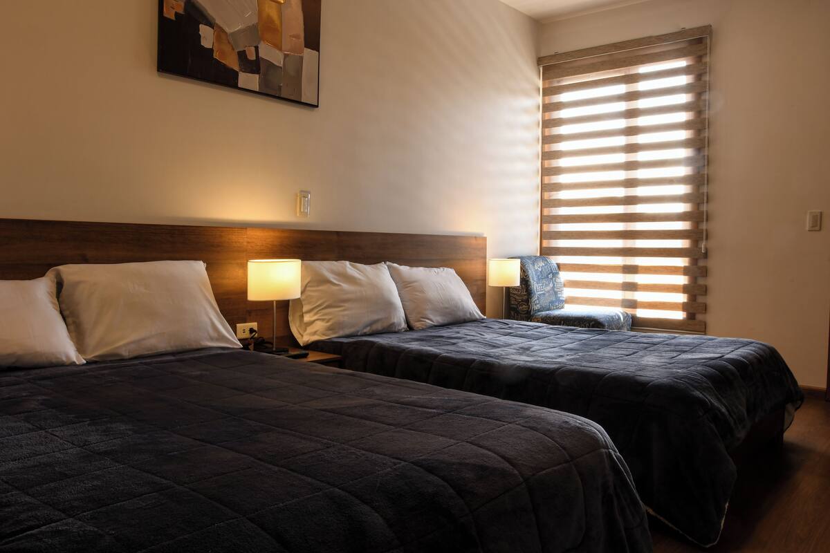 A hotel room with two beds, each with a headboard, bedside lamps, and a window with blinds.