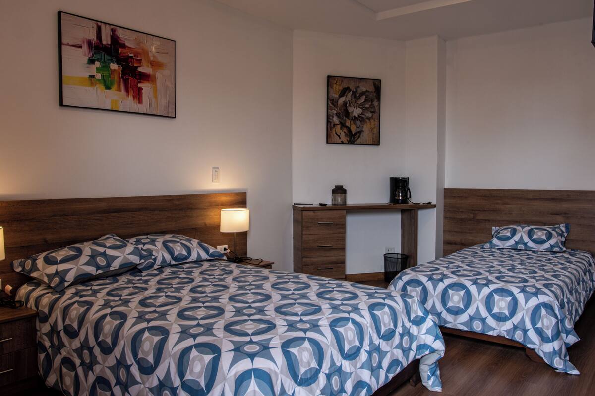A hotel room with two beds, a wooden headboard, a nightstand, a lamp, a coffee maker, and a shelf with a decorative item.