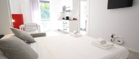 A modern hotel room with a large bed, a flat-screen TV, a small kitchenette, and a balcony with a red chair.