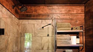 Baño de cabaña de madera con mampara de vidrio, baldosas de mármol y un estante con toallas y artículos de aseo.
