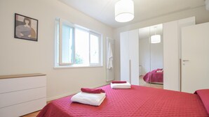 Een slaapkamer met een groot bed, een raam, een radiator en een deur naar een andere kamer.