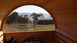 Una cabaña de madera con un ventanal arqueado grande que da a un espacio verde con un banco, una construcción pequeña y montañas al fondo.