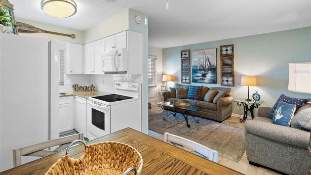 A modern kitchen with white cabinets, a built-in microwave, and a stove. There is a dining area with a wooden table and chairs, and a living room with a sofa and a coffee table. - Waves 4, poolside condo, 5 min walk to beach and entertainment (St. Pete Beach)