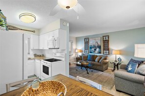 Una cocina moderna con armarios blancos, microondas integrado y estufa. Hay una zona de comedor con mesa y sillas de madera, y una sala de estar con sofá y mesa de centro.