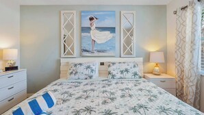A bedroom with a bed, two nightstands, a digital clock, and a painting of a woman by the sea. - Waves 4, poolside condo, 5 min walk to beach and entertainment (St. Pete Beach)