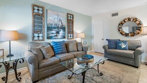 A living room with two grey sofas, a glass coffee table, and decorative items including a sailboat painting and a mirror. - Waves 4, poolside condo, 5 min walk to beach and entertainment (St. Pete Beach)
