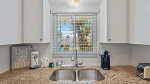 A kitchen with white cabinets, a double sink, a window with blinds, and a countertop with various items. - Waves 4, poolside condo, 5 min walk to beach and entertainment (St. Pete Beach)