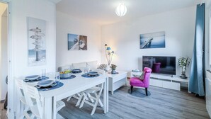 Un salon moderne avec un coin repas, comprenant une table à manger blanche, des chaises, un fauteuil rose, un téléviseur et des œuvres d’art accrochées aux murs.